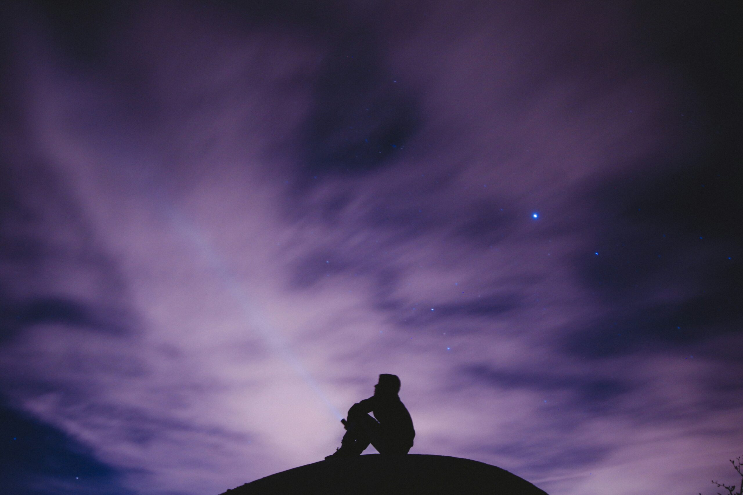 Silhouette di una persona seduta in contemplazione sotto un cielo viola stellato, simbolo di sogno, introspezione e spiritualità.